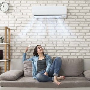 Eine Junge Frau sitzt in einem Raum auf der Couch mit der Fernbedienung für die Klimaanlage die an der Wand über Ihr ist. Aus der Klimaanlage kommt kühle Luft, und die Frau atmet Tief und lächelt.