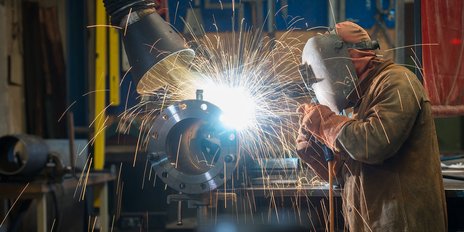 Ein Schweißer arbeitet konzentriert an einem Metallstück, während Funken in alle Richtungen sprühen und eine helle Lichtquelle entsteht.