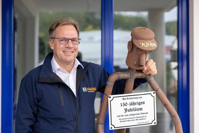 Ein lächelnder Mann in einer blauen Jacke steht neben einer rostigen Figur mit einer Jubiläumstafel in der Hand.